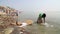 Man washing laundry on shore of Ganges, with city in the background.