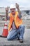 Man wearing hard hat and safety vest kneeling next to traffic cone gesturing with his hands