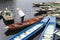 Man wearing white shirt, conical hat with his paddle stand on red empty rowing boat with many boats stop over the river.