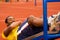 Man wearing yellow shirt and blue shorts lying down on wooden training plank doing situps smiling, orange athletic