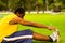 Man wearing yellow shirt and blue shorts sitting down on green grass stretching by touching shoes with hands, training