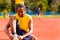 Man wearing yellow shirt and blue shorts sitting down tying shoe laces at outdoors training facility with orange