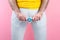 A man in white jeans, legs spread, holds a condom at the level of the genitals. Hands close-up. Pink background. Concept of
