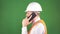 A man in a white protective helmet and signal vest talking on the phone at a construction site, green background