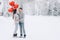 A man and a woman are hugging in a snowy park. A woman holds in her hands heart-shaped balloons filled with helium