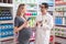 Man and woman pharmacist and pregnant client using smartphone speaking at pharmacy