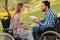 A man and a woman in wheelchairs met in the park. They exchange gifts and smile at each other.