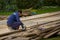 Man worker in blue work clothes handles wooden planks with grinding machine on background of green bushes and lawn