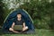 Man working on laptop in tent in nature. Young freelancer sitting in camp. Relaxing in camping site in forest, meadow. Remote work