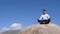 Man in yoga lotus pose meditating on stony mountain peak, blue sky background