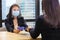 Manager from HR department wearing facial mask is interviewing new applicant who is handing her resume during social distancing