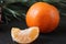 Mandarin whole in the peel and a slice of fruit lie side by side on a black background next to a branch of spruce and a silver