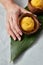 A mango ice cream sorbet in a coconut shell is held by a female hand on a palm leaf on a gray concrete background. Top