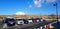 Many car parked on public parking on street with high voltage electrical tower, wire, blue sky and Fuji mountain background.