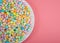 Many colorful candy jelly beans on porcelain plate laying on light pink surface