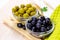 Many green and black marinated whole olives in the glass bowl on light wooden board background in the kitchen.