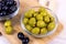 Many green marinated whole olives in the glass bowl on light wooden board background in the kitchen.