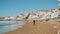 Many people relaxing on the beach in small village, ocean background. Boys playing with pink ball, gulls fly over