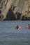 Many surfers sit and lie on their surfboards in front of green overgrown rocks on a beach in northern Spain