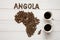 Map of the Angola made of roasted coffee beans laying on white wooden textured background with two cups of coffee