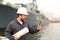 Marine engineer in helmet holding VHF walkie talkie and papers near vessel in background.