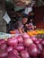 A market vendor sells red onions and other produce, surrounded by bustling shoppers and the enticing aroma of fresh vegetables.