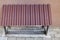 Maroon corrugated metal sunshade visor attached to iron posts embedded in cement floor above basement window on brown wall.