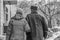 Married couple of pensioners walking arm-in-arm in the street with houses and trees and dirty snow in winter and an elderly woman