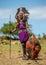 Masai warrior is standing in traditional clothing in a warrior`s headdress with spear and shield against the backdrop of savannah.