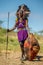 Masai warrior is standing in traditional clothing in a warrior`s headdress with spear and shield against the backdrop of savannah.