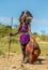 Masai warrior is standing in traditional clothing in a warrior`s headdress with spear and shield against the backdrop of savannah.