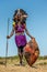 Masai warrior is standing in traditional clothing in a warrior`s headdress with spear and shield against the backdrop of savannah.