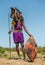 Masai warrior is standing in traditional clothing in a warrior`s headdress with spear and shield against the backdrop of savannah.