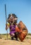 Masai warrior is standing in traditional clothing in a warrior`s headdress with spear and shield against the backdrop of savannah.