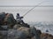 Masked angler in camouflage holding fishing rod in hands sitting on rocks coast in Aramoana Dunedin Otago New Zealand