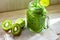 Mason jar mug with green vegetable and fruit smoothie, kiwi, lime, white wood table outdoors, sunlight fleck