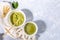 Matcha green tea cup and powder bowl, wooden spoon, bamboo whisk, fresh leaves on napkin on grey textured backdrop.