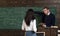 Math teacher pointing at green board. Professor giving explanations to brunette female student. Turn back girl in white