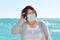 Mature asian woman with a surgical mask standing close to the sea and talking on her phone. Communication and leisure concept