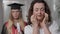 Mature Caucasian woman crying as blurred graduate daughter in gown and hat standing at background. Portrait of worried