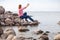 Mature Caucasian woman using smartphone for taking selfie while sitting on rocky seashore, female dressed red and white shirt and