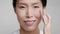 Mature Chinese Lady Applying Moisturizer Cream On Face, Gray Background