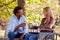 Mature Couple With Woman Sitting In Wheelchair Talking And Drinking Coffee In Park Together