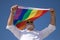 Mature, gray-haired gay man with beard, sunglasses and white shirt waving a gay pride flag in the wind under a blue sky. Concept