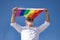 Mature, gray-haired gay man with beard, sunglasses and white shirt waving a gay pride flag in the wind under a blue sky. Concept