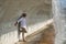 Mature, gray haired man with beard, sunglasses, white shirt and light brown pants, leaning against a concrete wall under a