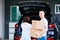 Mature happy Asian married couple carrying cardboard boxes from car trunk at new home.