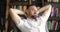 Mature man is dreaming resting in armchair in library with hands behind head.