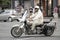 Mature man in light beige outfit riding a double seat motorbike with side saddle bags on city street traffic