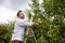 Mature man picking apples in orchard. Person stands on a ladder near tree and reaching for an apple